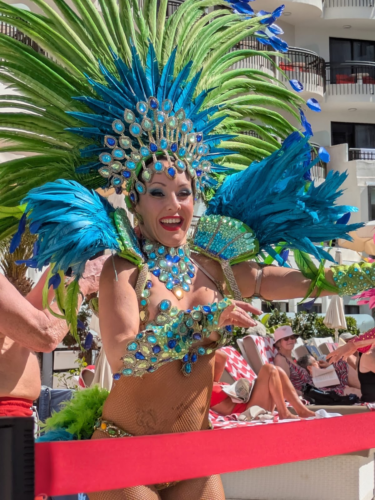 Costumed carnival dancer in a feathered headdress smiling beside a hotel pool