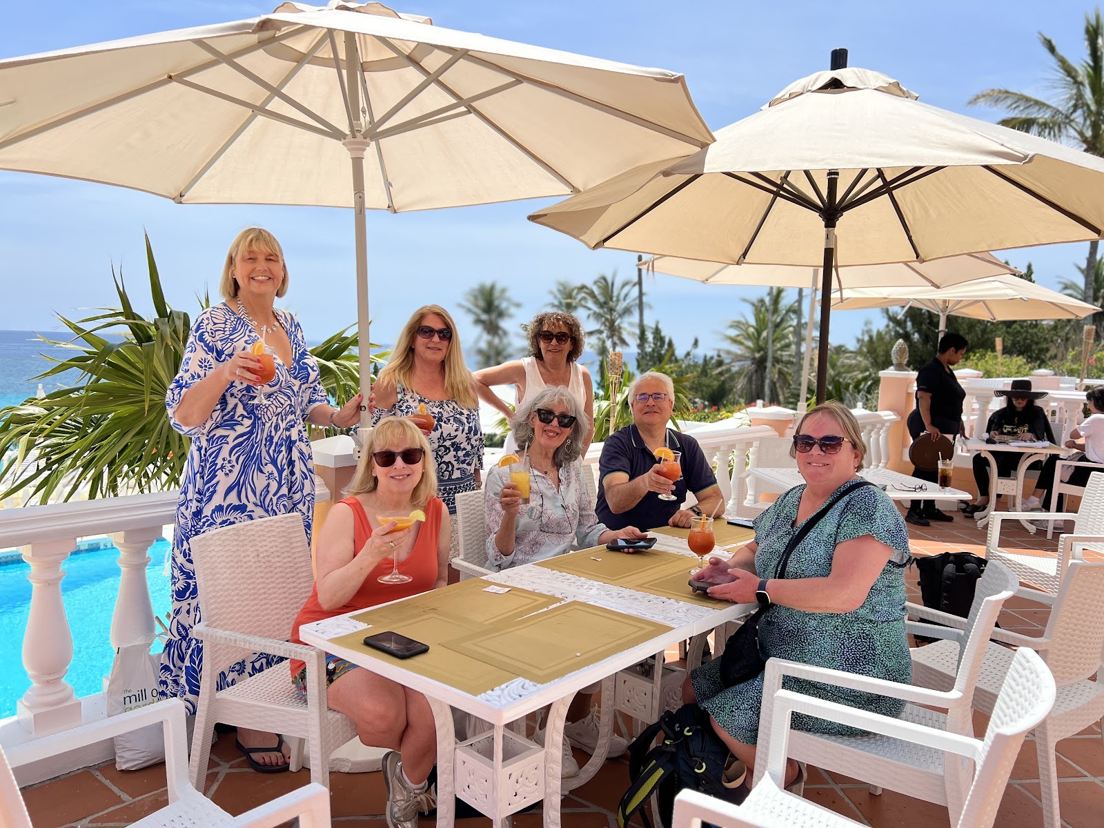 Six travellers with drinks at a poolside table under white parasols