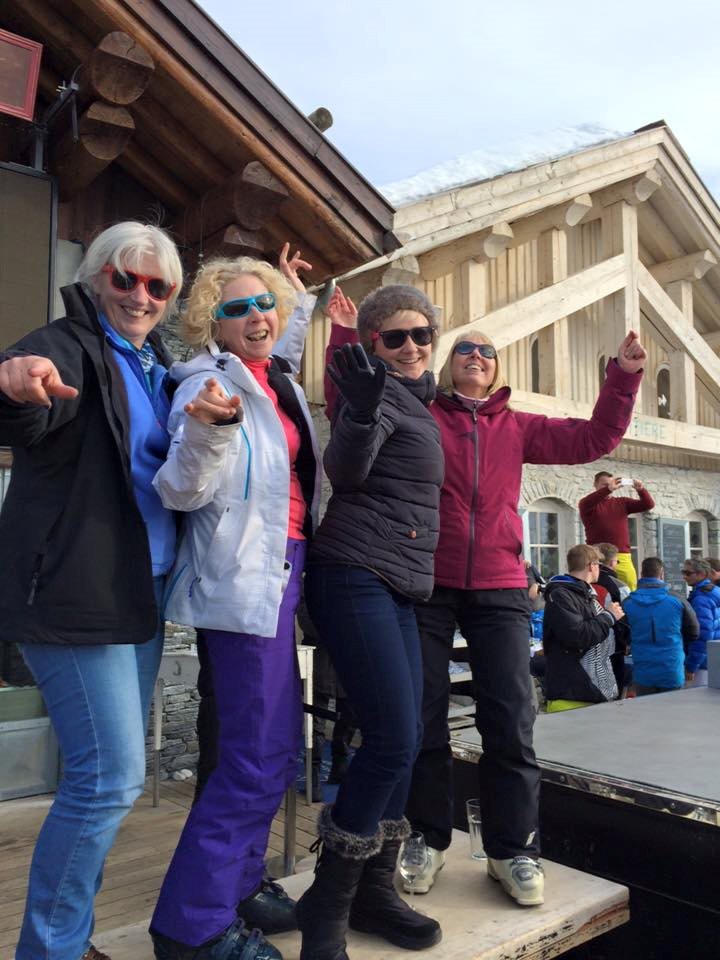 Four women in ski gear laughing and pointing at the camera outside a chalet