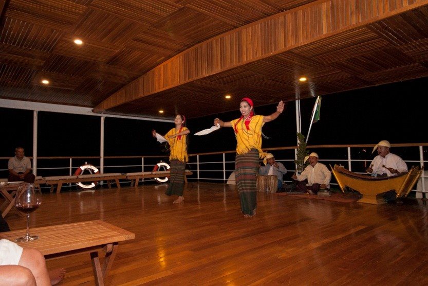 Two traditional dancers performing on a boat deck with musicians and seated guests
