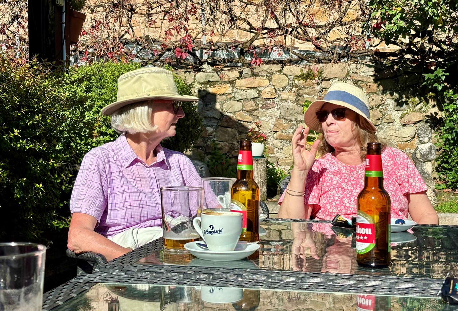 Two women sitting at a garden table with drinks, chatting in the sun