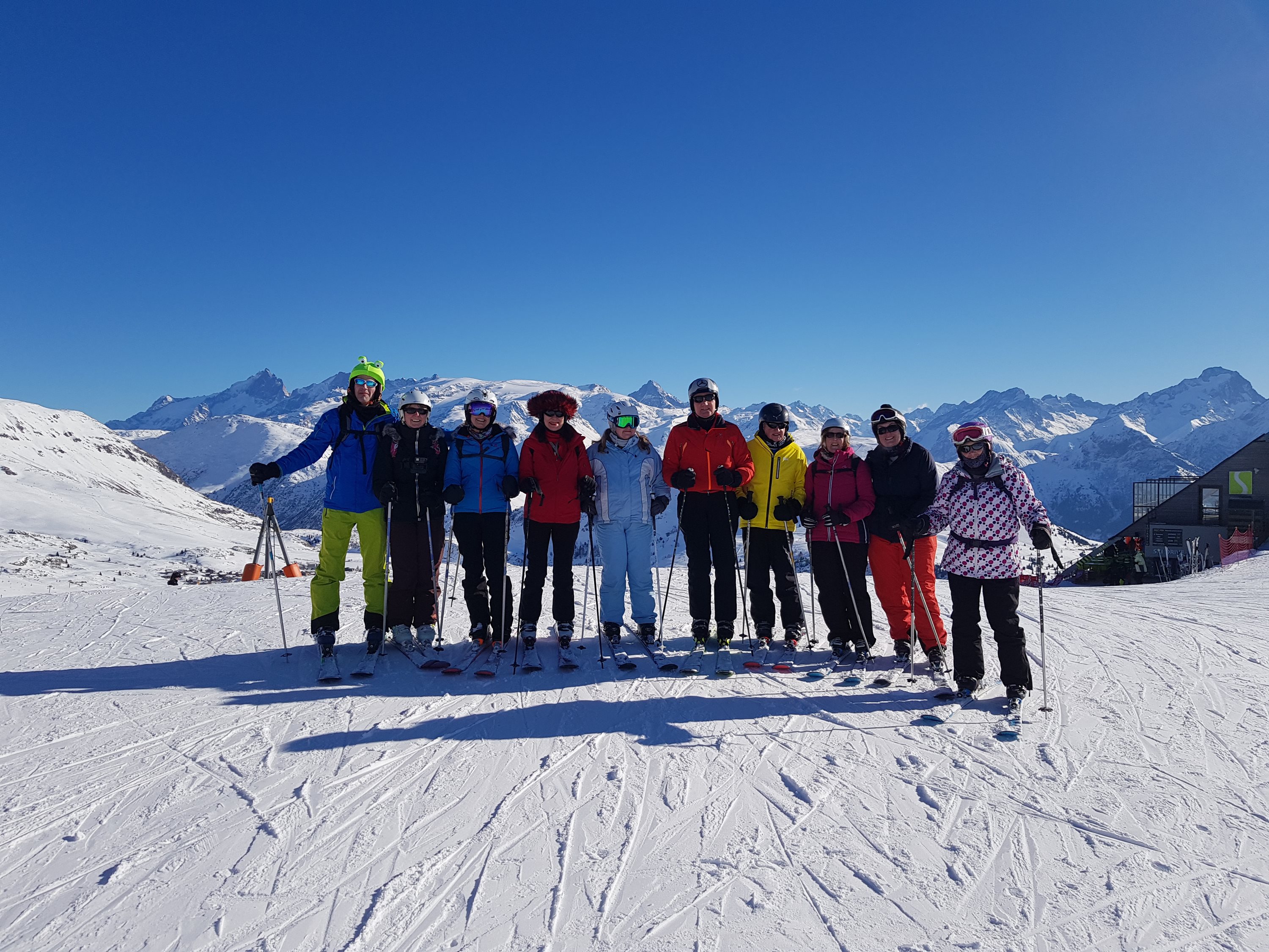 A line of skiers posing arm in arm on the piste with mountains behind them