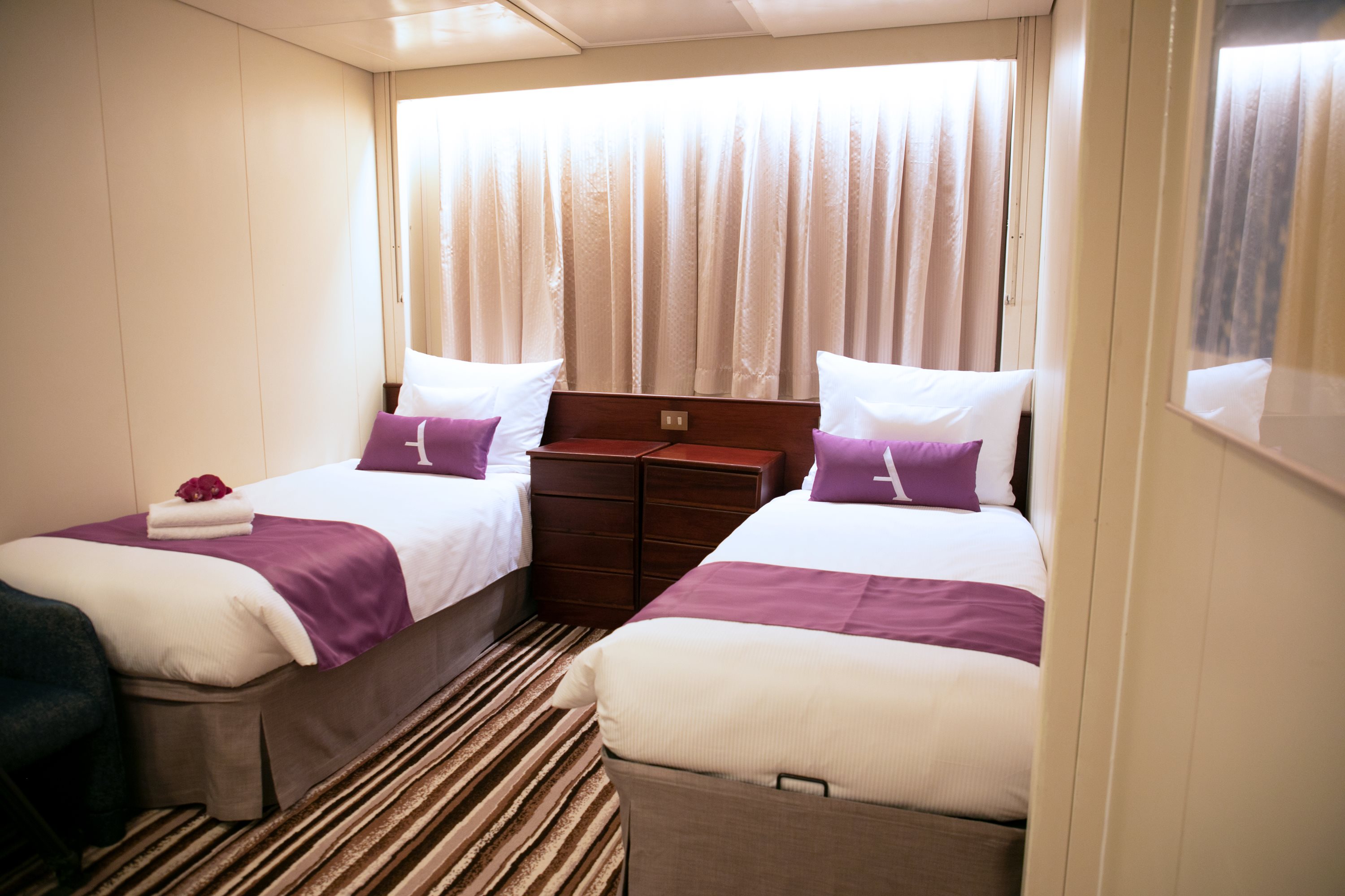 Twin-bedded cruise cabin with purple bed linen and a curtained window