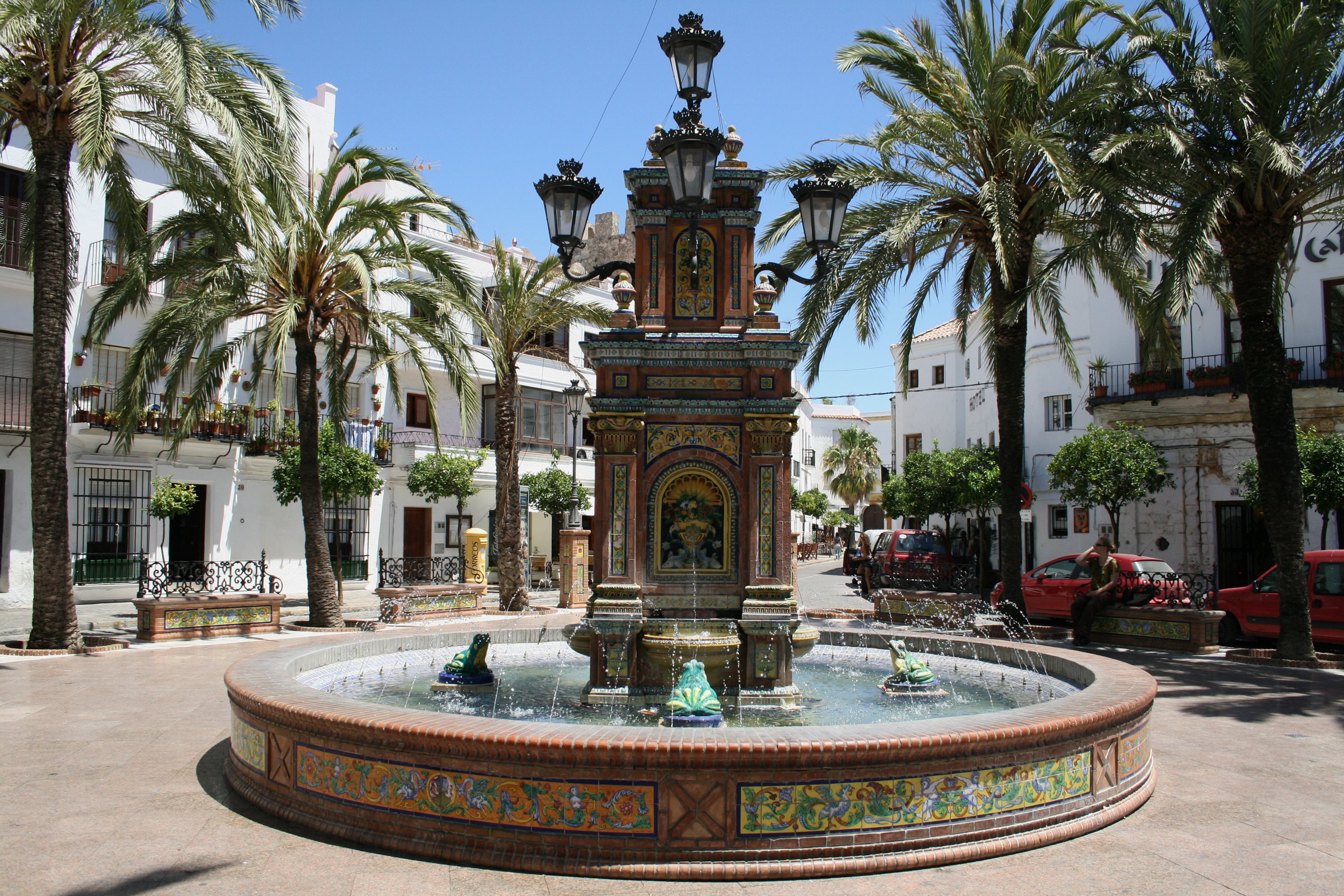An ornate tiled fountain in a plaza lined with palm trees and whitewashed buildings