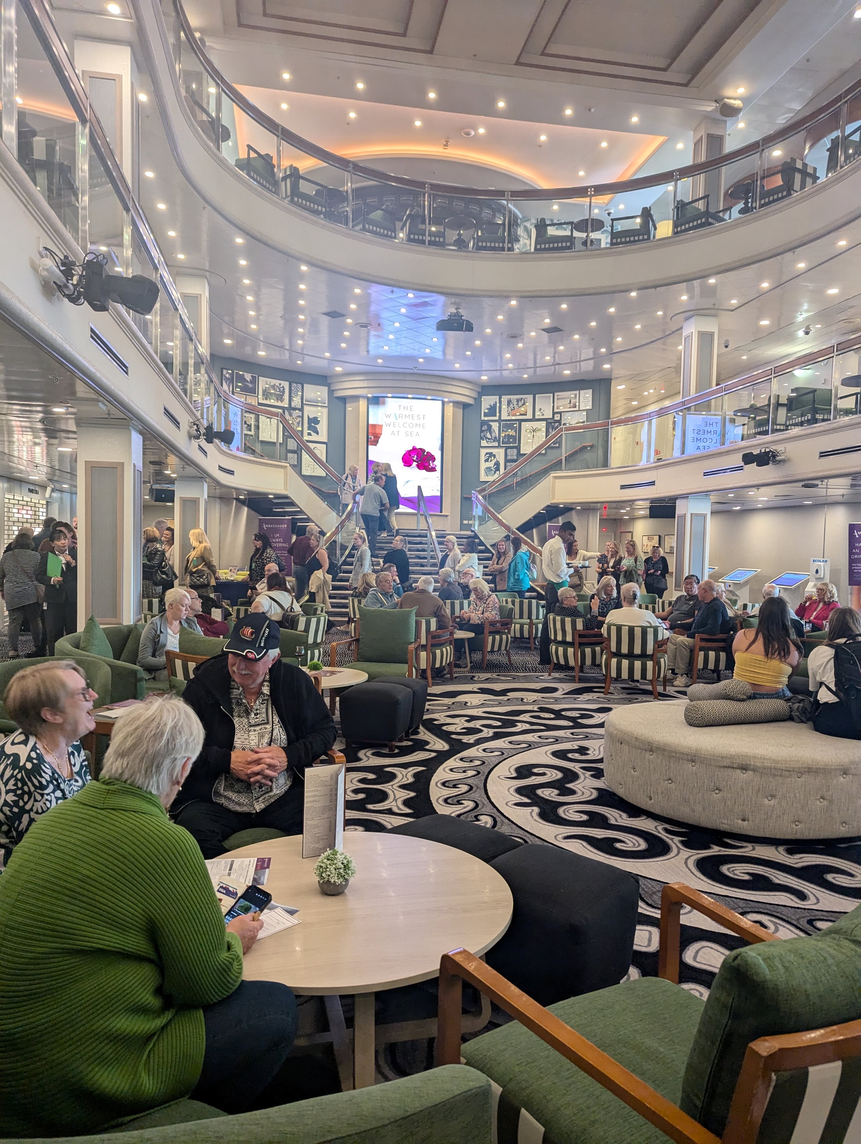 Two women chatting at a table in a busy ship atrium lounge with other passengers around