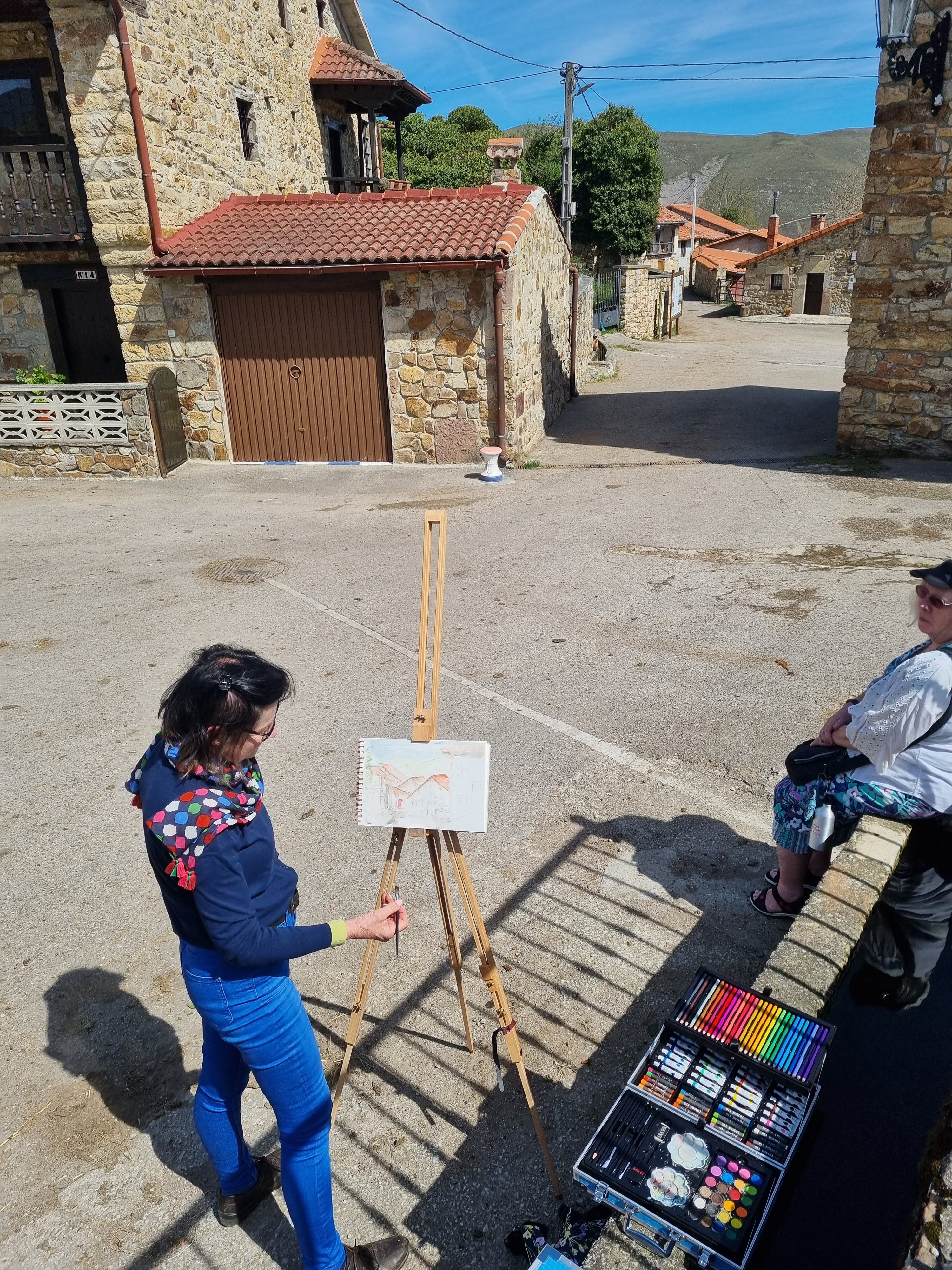 Woman painting at an outdoor easel in a village street while another looks on