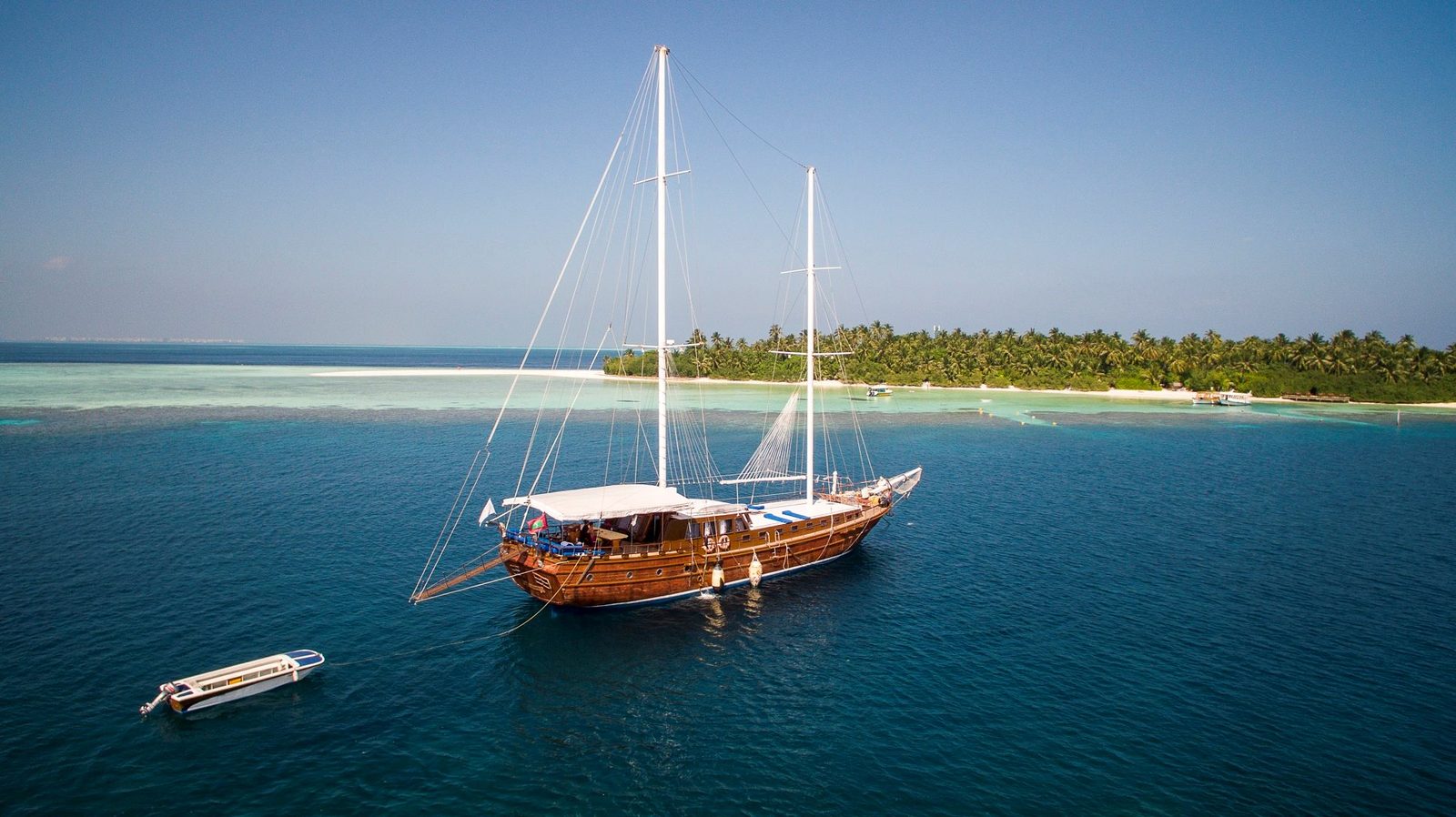 Traditional wooden sailing boat anchored near a Maldivian island with a tender alongside