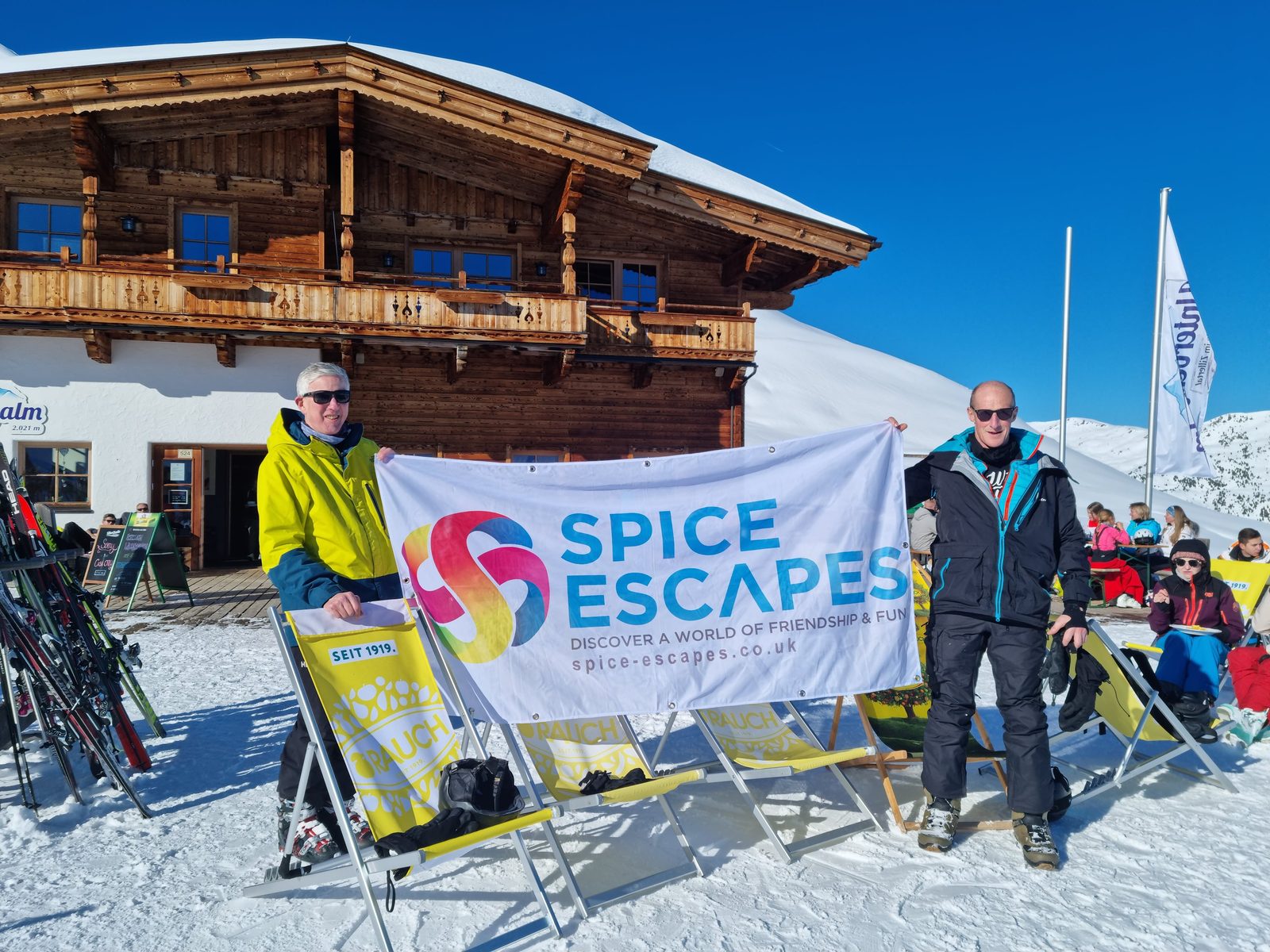 Two men posing with a Spice Escapes banner outside a mountain chalet