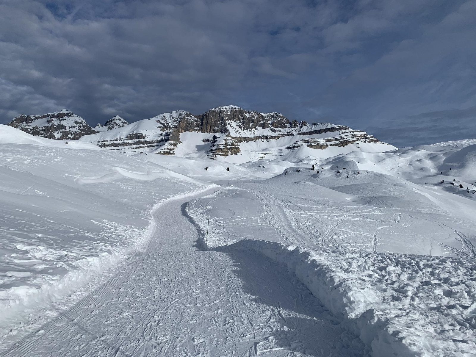 A groomed piste winding towards rocky peaks under a cloudy sky
