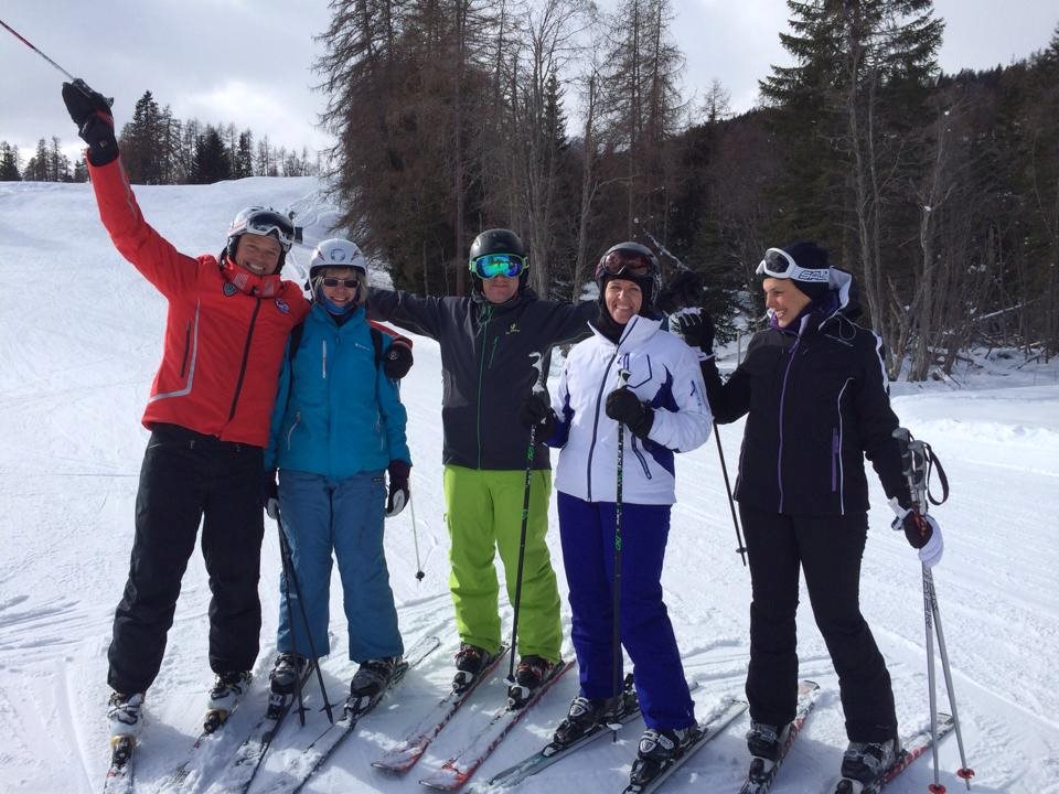 Five skiers standing side by side on the piste, poles raised above their heads