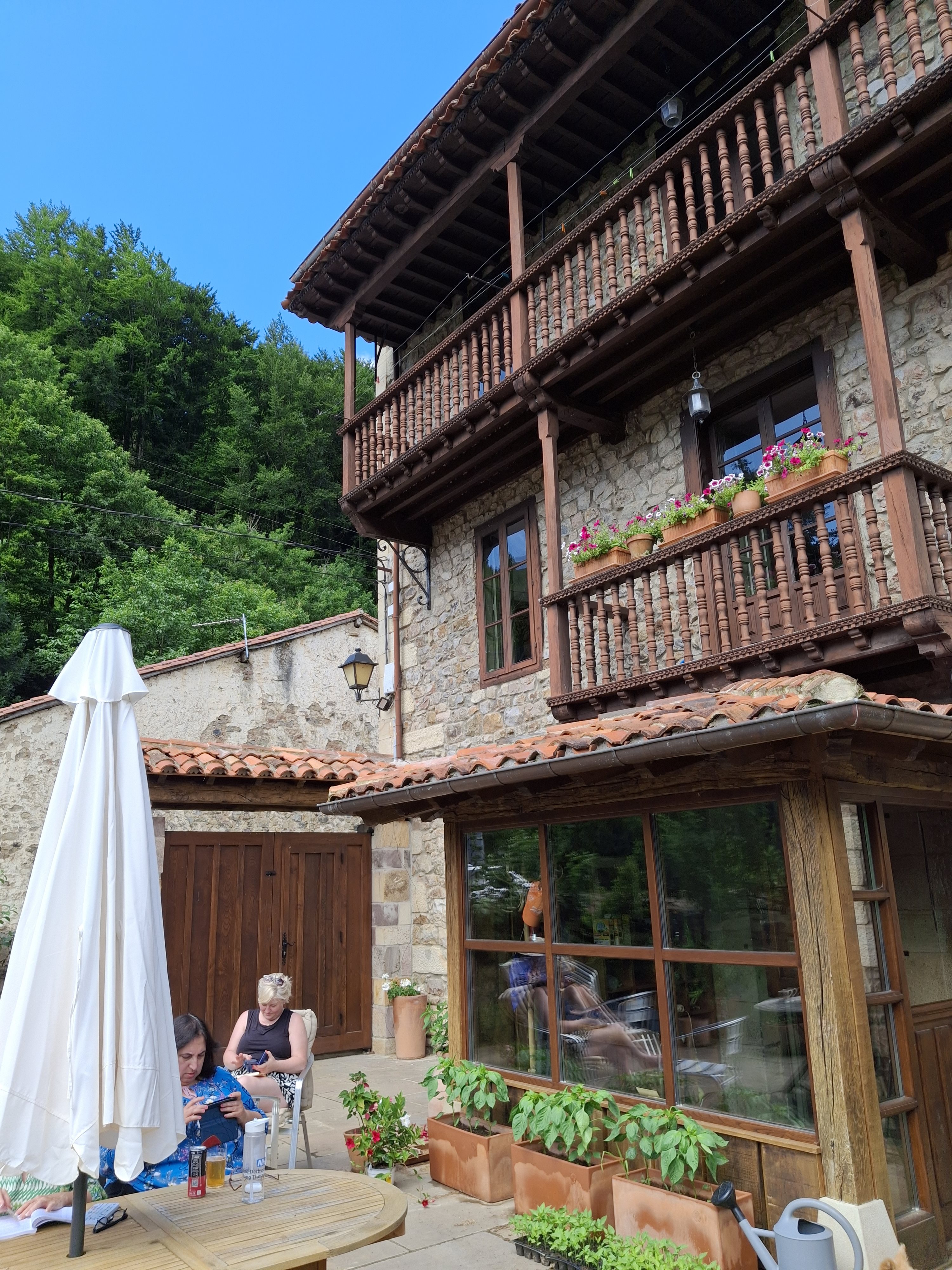 Two women sitting at an outdoor table below a traditional balconied stone house