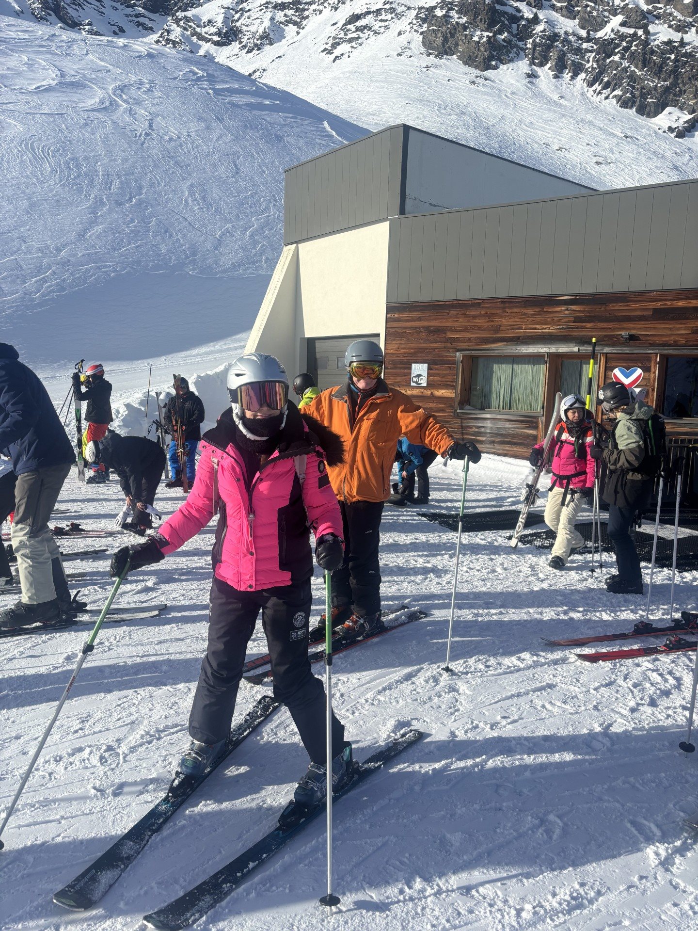 A skier in a pink jacket poses on the slope with others and a lodge behind