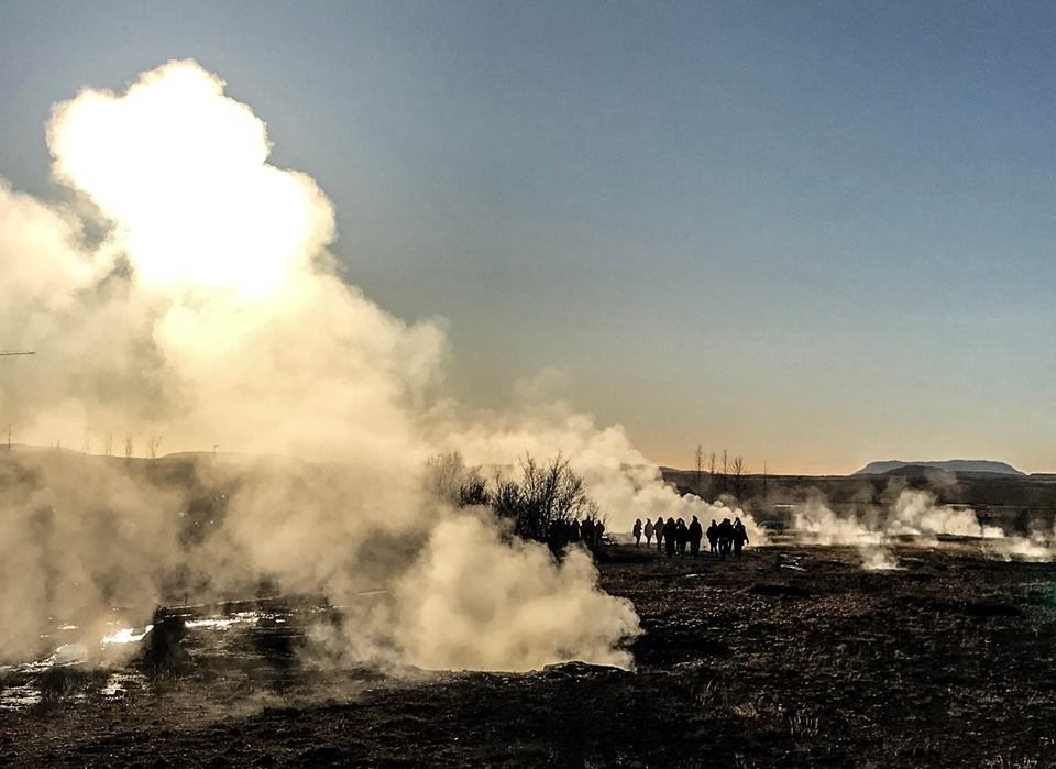 A group of visitors watching steam rise from a geothermal area at sunset