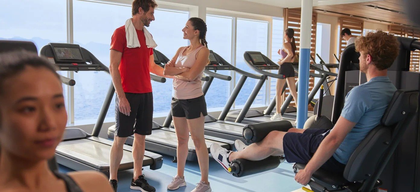 Two guests chatting in a ship's gym while others use treadmills and weights