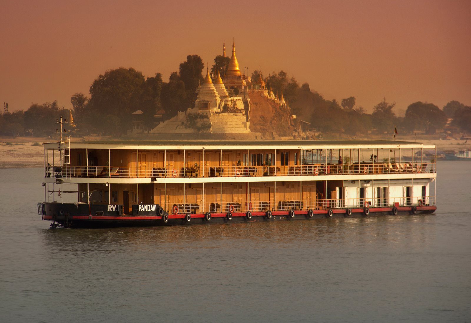 A river cruise boat passing a golden temple-topped hill at sunset