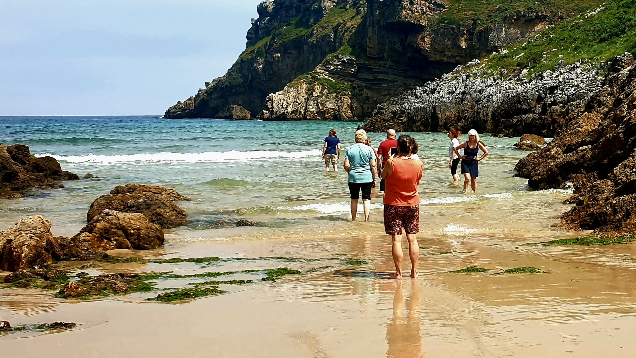 A small group wading into the sea from a sandy beach between cliffs